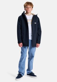 Marineblauer wasserdichter Mantel mit einer gefütterten Kapuze aus Fell, kombiniert mit einem weißen T-Shirt und hellblauen Jeans, präsentiert von einem aufrecht stehenden Model.
