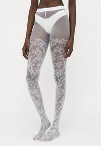 Lace-mønstret strømpebukser i gennemsigtig grå med blomsterdesign. Har en solid hvid talje og åbne tæer. Blød, strækbar tekstur.