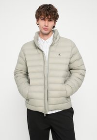 Casaco acolchoado cinza claro com fecho de correr frontal, gola alta e bolsos laterais. Apresenta costuras horizontais e um pequeno logótipo no peito.