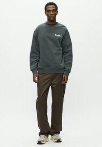 Mørkegrøn sweatshirt med ribstrik i ærmer og bund, med hvidt logo. Kombineret med løse brune bukser og lyse sneakers.