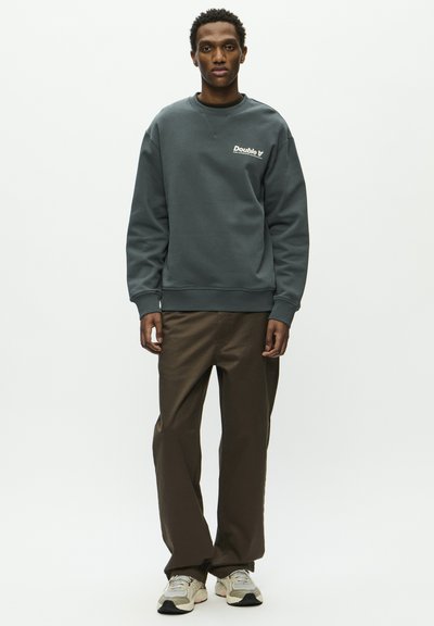 Sweatshirt vert foncé avec des poignets et un ourlet côtelés, arborant un logo blanc. Associé à un pantalon large marron et des baskets de couleur claire.