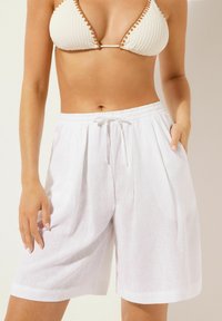 Shorts en lin blanc avec une coupe décontractée, taille élastique, détails à cordon de serrage et poches latérales. Texture lisse avec un léger aspect transparent.