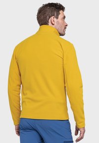 Gelbe Fleecejacke mit hohem Kragen, langen Ärmeln und glatter Textur, getragen über blauen Hosen. Keine sichtbaren Nähte oder Logos.