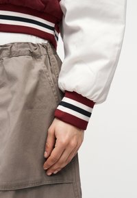 Krupan plan ruke koja počiva pored bež hlača s elastičnim pojasom i bijelog rukava jakne s crveno-bijelo-crnim prugastim rubom.