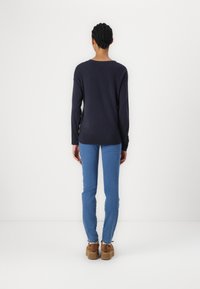 Kvinna med flätat hår iklädd en marinblå tröja, blå jeans och bruna platåskor, stående vänd mot en vit vägg.