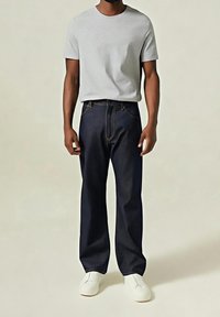 Homme portant un t-shirt gris clair à manches courtes uni, un jean ample de couleur foncée et des chaussures blanches sans lacets, debout devant un fond uni.