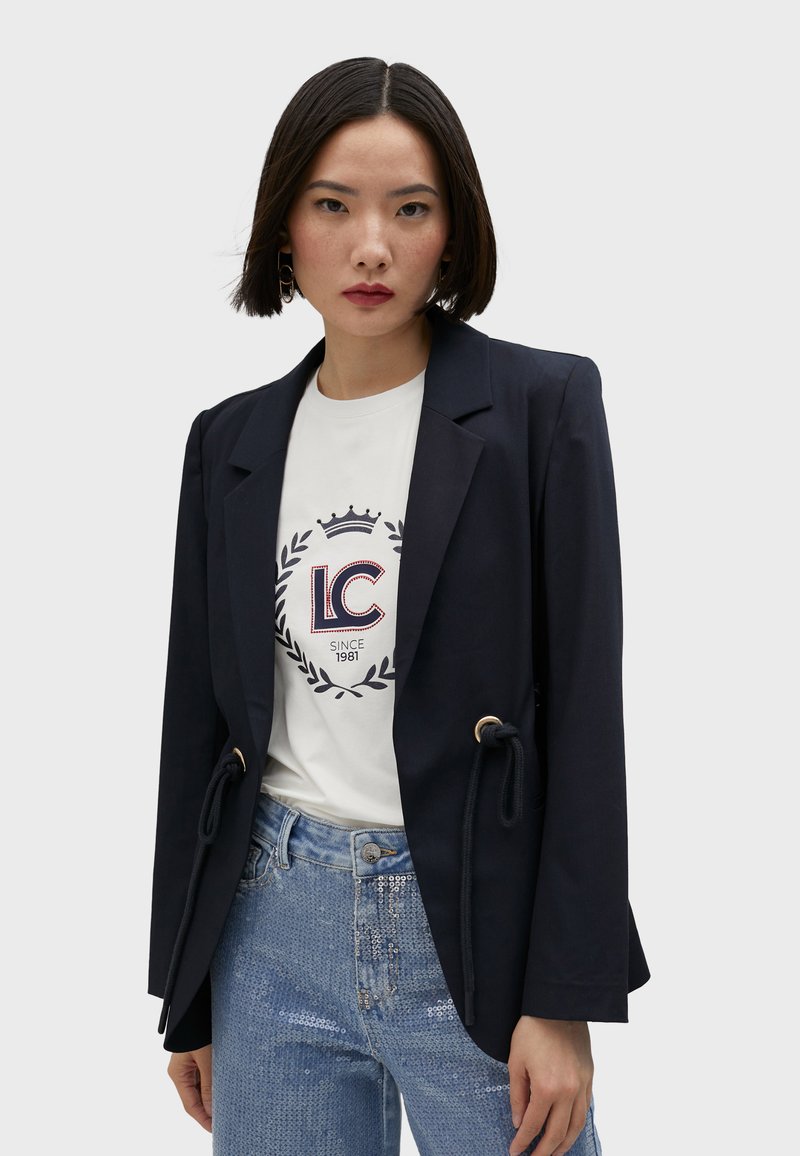 Navy blazer with a tailored fit, tie details, and gold accents over a white graphic t-shirt, paired with shiny sequined light blue jeans.