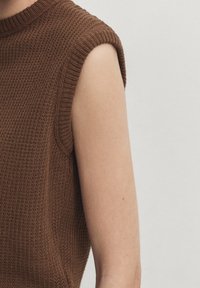Bruine gebreide mouwloze top met ribbeltextuur en een hoge halslijn, met nadruk op de schouder- en armdetails.