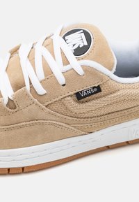 Sneakers Vans bej cu material din suede, cu șireturi albe și un patch logo negru. Prezintă un panou lateral texturat și o talpă din cauciuc de tip gum.