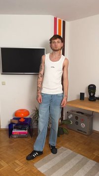 Λευκό αμάνικο μπλουζάκι με λογότυπο, ανοιχτό μπλε τζιν με ίσια γραμμή, μαύρα αθλητικά παπούτσια με τρεις λευκές ρίγες, τατουάζ ορατά στα χέρια.