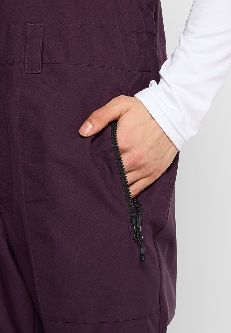 Fioletowe ogrodniczki z boczną kieszenią na zamek błyskawiczny. Materiał wydaje się gładki, z dopasowanym fasonem w okolicy talii i nóg.