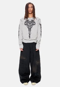 Νεαρός με μακριές dreadlocks φοράει γκρι μακρυμάνικο πουκάμισο με μαύρο σχέδιο φυλετικού κρανίου και άνετο μαύρο παντελόνι cargo με αθλητικά παπούτσια.