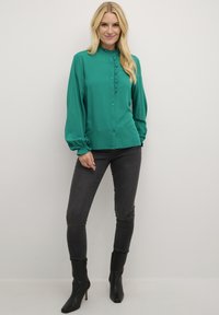Blusa verde a maniche lunghe in tessuto liscio, con frontale arricciato, patta con bottoni e colletto arrotondato. Abbinata a jeans scuri.