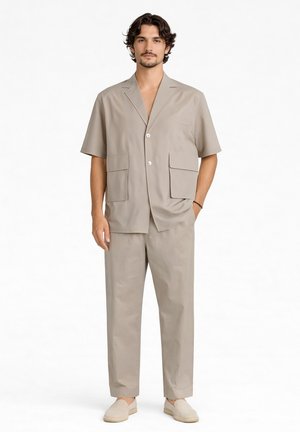 Uomo in piedi rivolto di fronte che indossa un blazer beige a maniche corte, pantaloni abbinati e scarpe senza lacci su sfondo bianco.