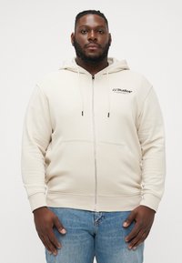 Beige zip-up hoodie gemaakt van zachte stof, met een voorzak, verstelbare trekkoordkap en het JJ Studios-logo op de linkerborst.