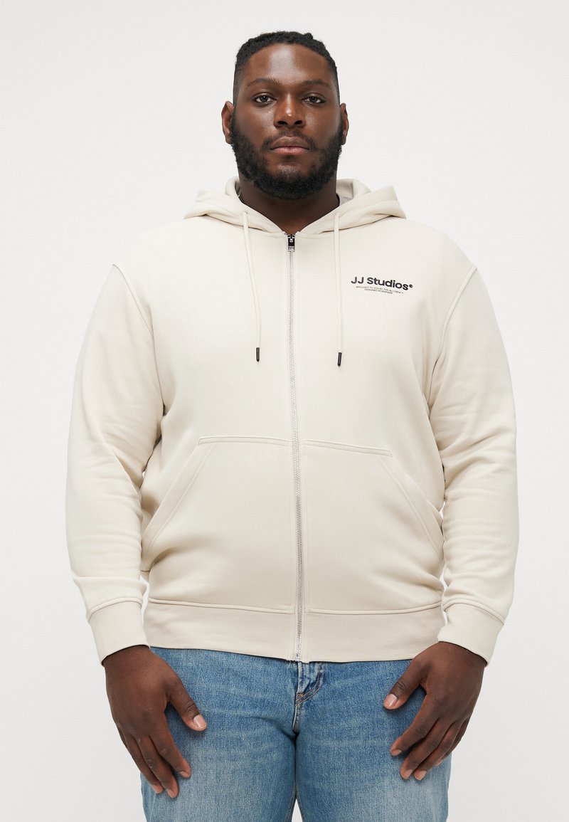 Beige zip-up hoodie gemaakt van zachte stof, met een voorzak, verstelbare trekkoordkap en het JJ Studios-logo op de linkerborst.