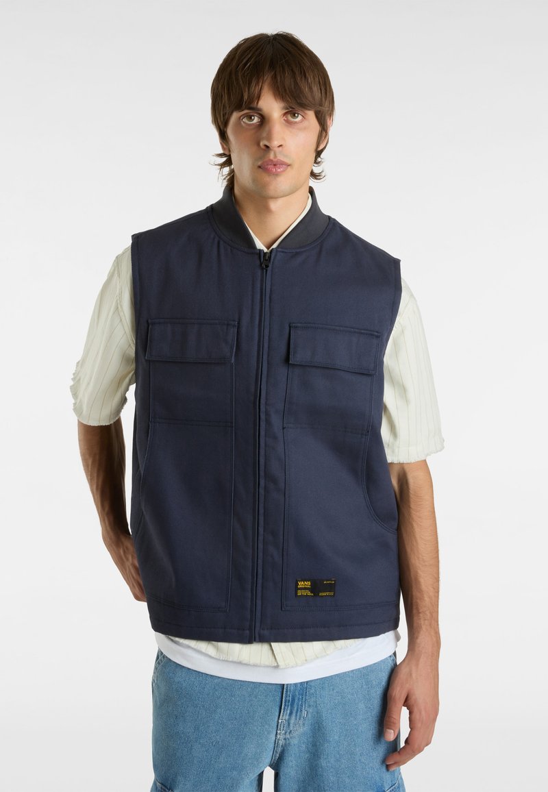 Gillet senza maniche blu navy con zip frontale, due tasche sul petto e un'etichetta vicino all'orlo. Indossato sopra una camicia a righe leggera e jeans casual.