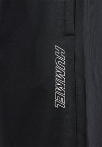 Nærbillede af sort stof med vertikalt hvidt HUMMEL-logo trykt, der viser søm og teksturdetaljer.