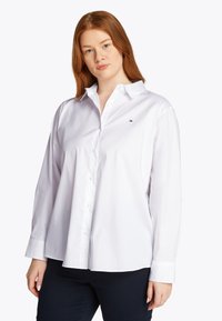 Λευκό πουκάμισο με κουμπιά, μακριά μανίκια, κλασικό γιακά, λείο βαμβακερό ύφασμα, μικρή λεπτομέρεια λογότυπου στο στήθος, ίσια γραμ μή, μινιμαλιστικός σχεδιασμός.