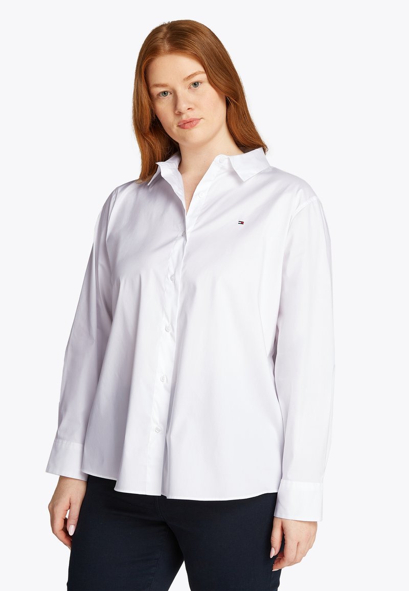 Λευκό πουκάμισο με κουμπιά, μακριά μανίκια, κλασικό γιακά, λείο βαμβακερό ύφασμα, μικρή λεπτομέρεια λογότυπου στο στήθος, ίσια γραμμή, μινιμαλιστικός σχεδιασμός.