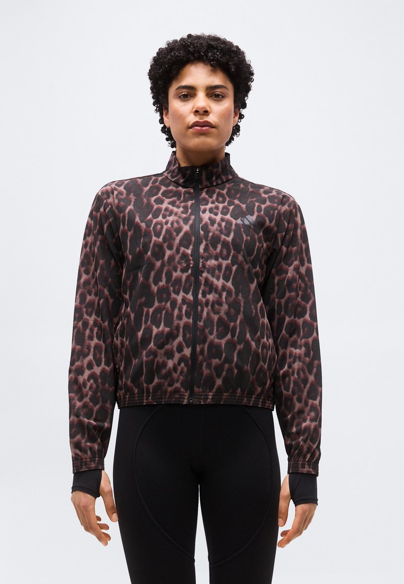 Personne portant une veste à manches longues zippée à imprimé léopard avec des trous pour les pouces, associée à un pantalon noir ajusté, sur un fond uni.