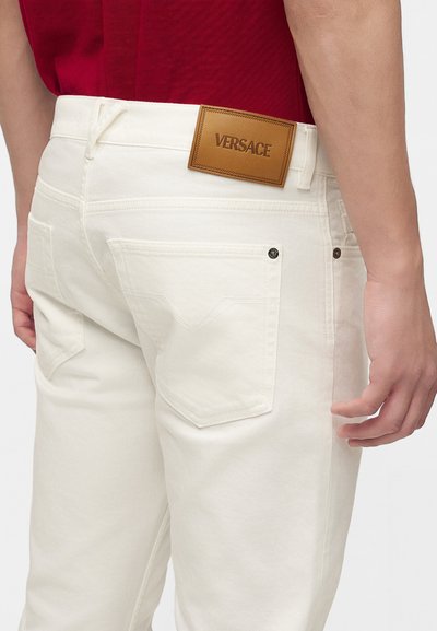 Personne portant un jean blanc avec des poches arrière et une étiquette en cuir marron de la marque Versace au-dessus de la poche droite, associé à une chemise rouge.