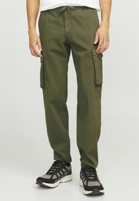 Uomo che indossa pantaloni cargo verde oliva con tasche a patta sui lati e scarpe sportive nere e grigie, in piedi contro uno sfondo semplice.