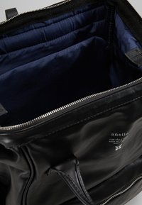 Bolsa de cuero negra con una textura suave, parte superior con cremallera y forro interior espacioso en color azul marino. Cuenta con una etiqueta de marca y asas resistentes.