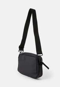Čierna crossbody taška vyrobená z hladkého materiálu, s uzatváraním na zips, nastaviteľným popruhom a obdĺžnikovým tvarom so zaoblenými hranami.