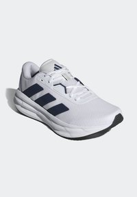 Chaussure de course blanche avec des rayures noires, des lacets blancs et une semelle amortie, présentée légèrement en angle sur un fond gris clair.