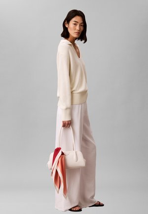 Mujer con suéter de punto color crema y pantalones anchos claros sostiene bolso blanco con pañuelo rojo-naranja, de pie contra fondo gris liso.