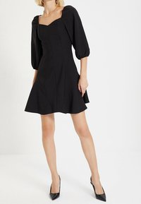 Vestido negro con mangas abullonadas, escote cuadrado, corsé ajustado y falda acampanada. Tejido suave con líneas limpias, combinado con tacones altos negros.