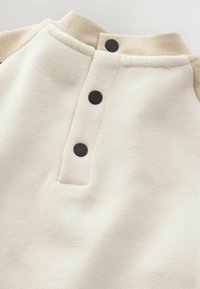 Sweatshirt couleur crème avec un col rond, des boutons noirs et des accents beiges sur les épaules. Texture douce avec un design décontracté.