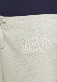Un pantalon de survêtement gris clair présente une texture douce, une taille avec cordon de serrage et un logo "GAP" brodé en blanc. Le haut est en tissu côtelé bleu marine.
