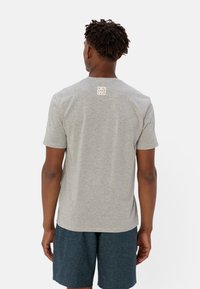 T-shirt grigio a maniche corte con collo a coste, con un piccolo etichetta rettangolare che recita "RECUPERATO" sul retro. Indossato con pantaloni corti blu navy.