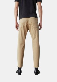 Pantaloni in cotone beige con una vestibilità su misura, dotati di tasche posteriori e un sottile motivo di cucitura. Indossati con stivaletti neri alla caviglia.