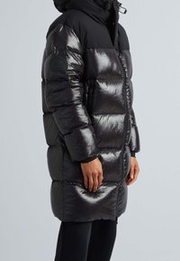 Svart vadderad vinterjacka med en glänsande yta, dragkedjestängning och huva. Har quiltade sektioner och en logotyp-patch på ärmen.