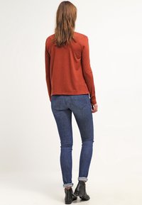 Rdzawoczerwony top z długim rękawem o luźnym kroju, w zestawieniu z ciemnoniebieskimi obcisłymi dżinsami i czarnymi botkami до kostek, sfotografowany od tyłu.