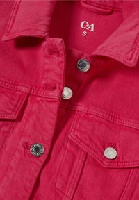 Červená džínsová bunda s límcem, stříbrnými druky a dvěma náprsními kapsami. Tkanina má texturovaný povrch a je složena k vystavení.