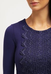 Marinblå klänning med långa ärmar som har en vågig spetsöverdel kring halslinningen, som visar cirkulära blommönster i texturerad detaljering.