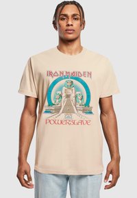 Beige bomull T-skjorte med et Iron Maiden-trykk. Designet inkluderer egyptiske motiver og fet rød tekst som sier "Powerslave."