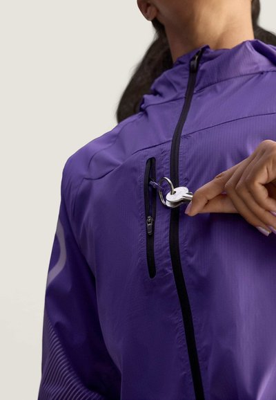 Chaqueta morada con cremallera, hecha de material ligero, que cuenta con un bolsillo en el pecho con cremallera, acentos negros y un acabado texturizado con rayas diagonales.
