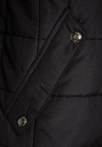 Primo piano di una giacca imbottita nera, con un tessuto testurizzato, trapuntatura diagonale e un bottone a pressione metallico sul lato sinistro.