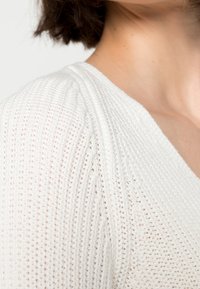 Biały dziergany sweter z teksturowanym wzorem, z głębokim dekoltem w serek i lekko bufkowatymi szwami na ramionach.