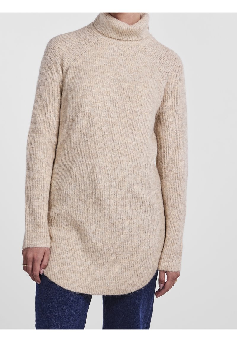 Beżowy prążkowany sweter z golfem o długich rękawach, zaokrąglonym kroju i miękkiej fakturze, zestawiony z ciemnoniebieskimi dżinsami.