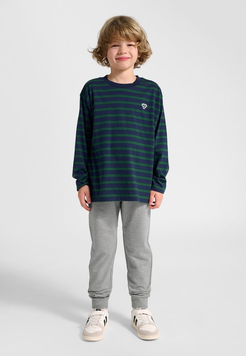 Kind lächelt, trägt ein marineblau-grün gestreiftes Langarmshirt, graue Jogginghose und weiße Turnschuhe vor einem einfarbigen hellen Hintergrund.