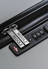 Close-up van koffer ritsen gegraveerd met "REDOLZ" en een combinatie slot met drie cijfers op een gestructureerd zilveren bagageoppervlak.