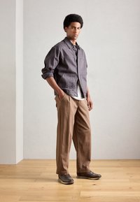 Hombre de pie en interiores vistiendo una chaqueta gris sobre una camisa blanca, pantalones sueltos marrones y zapatos casuales marrones oscuros, con las manos en los bolsillos.