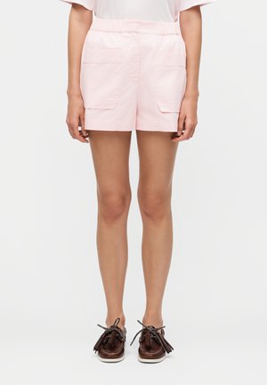 Nő világos rózsaszín rövidnadrágot visel két első zsebes fedéllel és barna bojtos mokaszin cipővel, egyszínű háttér előtt áll.