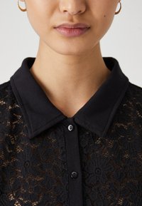 Svart blonde skjorte med knappelukking foran og rund krage. Intrikate blomsterdesign pryder stoffet, noe som gir det et teksturert utseende.